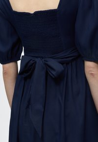 Πλάτη σκούρου μπλε φόρεματος με φουσκωτά μανίκια, σφηκοφωλιά τετράγωνης πλάτης και δεμένη φιόγκο στη μέση.
