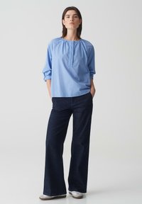 Blusa azzurro chiaro a maniche lunghe con scollatura arricciata, abbinata a pantaloni a gamba larga in denim scuro e scarpe grigie slip-on.