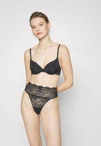 Soutien-gorge noir avec bonnets lisses et bretelles réglables, associé à un string en dentelle noire assorti, doté d'un motif floral et de bords festonnés.