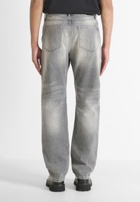 Persona vistiendo jeans de corte holgado en gris deslavado y zapatos negros gruesos, de pie con los brazos relajados contra un fondo blanco liso.
