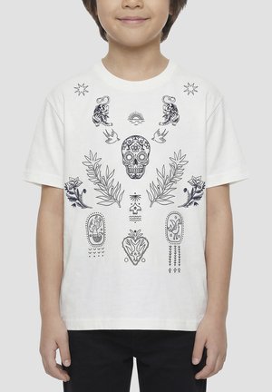 Bambino che indossa una maglietta bianca con disegni neri intricati di teschi, fiori, uccelli, tigri e cuori, in piedi davanti a uno sfondo semplice.