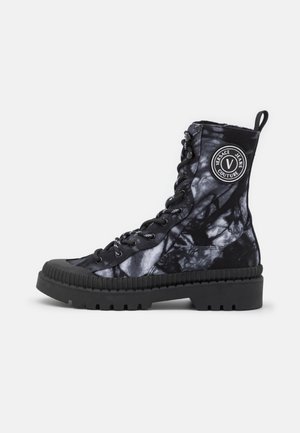 Czarno-szary botek z wzorem tie-dye, sznurowany, z grubą podeszwą, okrągłym noskiem, pętelką do łatwego zakładania oraz naszywką z logo "Versace Jeans Couture" po boku.