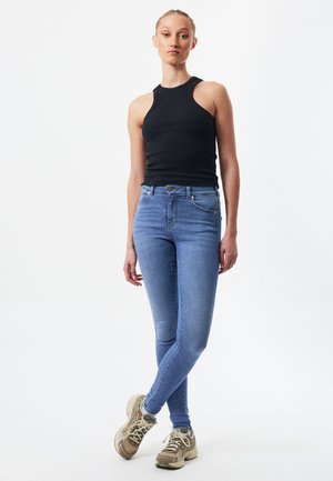 Top negro de canalé sin mangas, jeans ajustados azul claro y zapatillas beige con cordones; la modelo posa con una pierna cruzada, fondo neutro.
