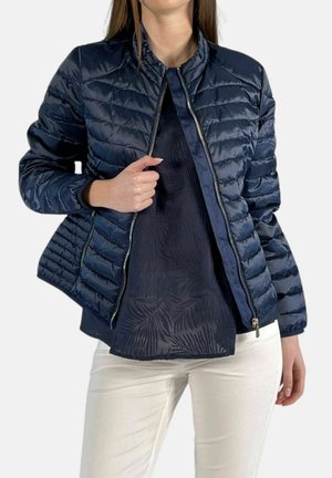 Donna che indossa pantaloni bianchi e una giacca trapuntata blu scuro sopra una maglia scura fantasia, aprendo parzialmente la giacca.