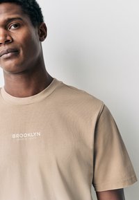 Krátke tričko s krátkym rukávom z hladkej látky s okrúhlym výstrihom. Na hrudi je biely nápis "BROOKLYN CEDAR ST. SOUTH".