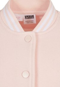 Giacca bomber rosa chiaro con colletto a strisce bianche, chiusura a scatto, tessuto morbido e etichetta interna che indica la taglia e le istruzioni per la cura.