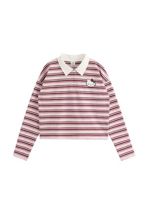 Ung kvinde med langt blondt hår iført en pink stribet langærmet skjorte med hvid krave og blå jeans, står med den ene hånd på hoften.