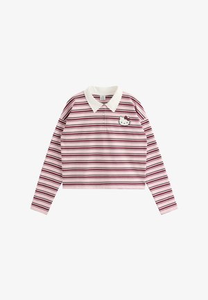 Ung kvinde med langt blondt hår iført en pink stribet langærmet skjorte med hvid krave og blå jeans, står med den ene hånd på hoften.