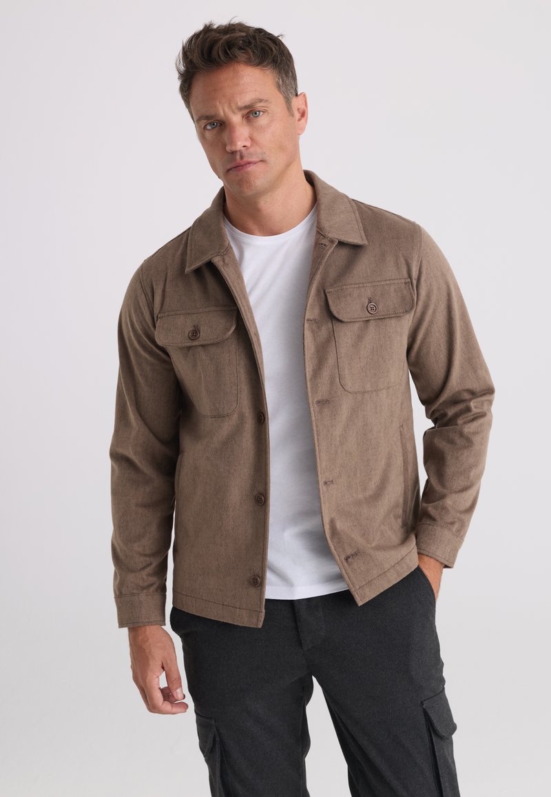 Homme portant une veste marron clair boutonnée par-dessus une chemise blanche et un pantalon cargo foncé, debout avec une main dans la poche devant un fond uni.