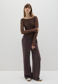 Top de manga larga marrón con encaje y escote amplio, combinado con pantalones sueltos de color púrpura oscuro y zapatos de suela blanca. Fondo de estudio.