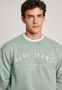Gaiši zaļš krekls no mīksta auduma ar apaļu kakla izgriezumu, ar drukātu tekstu "PEPE JEANS LONDON W11" baltā krāsā un ar teksturētu apdari.