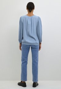 Blouse bleu clair à manches longues avec un tissu texturé, épaules froncées et col rond, associée à un jean bleu clair à jambes droites et des chaussures noires.