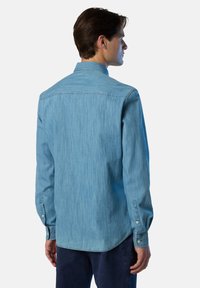 Camicia in denim azzurro chiaro con colletto a bottoni, maniche lunghe e polsini a bottoni. Presenta una texture leggermente sbiadita e un orlo curvo.