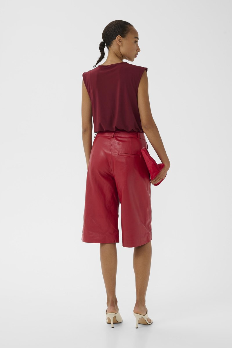 Kvinde med fletninger iført ærmeløs burgundy top, højtaljede røde skindshorts til knæet, beige hæle, der holder en rød clutch.