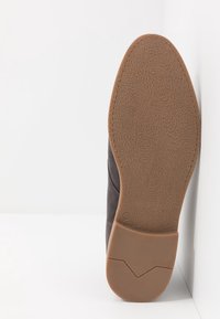 Chaussure en daim gris foncé avec une semelle en caoutchouc marron texturée, présentant un bout arrondi et un détail de couture subtil sur le côté. Taille indiquée : 42.
