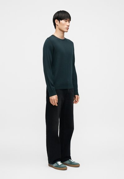 Pull vert foncé à manches longues, col rond, porté avec un pantalon large noir et des baskets bleu sarcelle à lacets blancs, debout sur un fond uni.