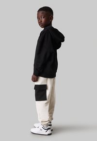 Zwarte hoodie gecombineerd met crèmekleurige joggers met een zwarte zijzak; witte sneakers met zwarte en groene accenten.