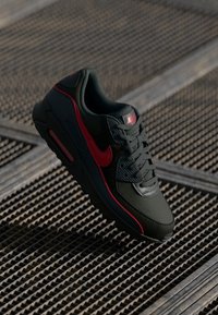 Crne i crvene Nike Air Max tenisice s vidljivom zračnom jastučićem, vezice vezane, odmaraju se na metalnoj rešetkastoj površini u prirodnom svjetlu.