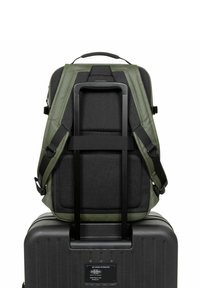 Mochila verde oscura con textura de malla y correas acolchadas, montada sobre una maleta negra con ruedas, que cuenta con un asa resistente y una superficie lisa.