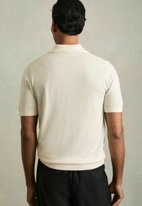 Polo cu mânecă scurtă de culoare crem, realizat dintr-un material neted, cu guler, mâneci ribbed și un croi confortabil, purtat cu pantaloni închiși la culoare.