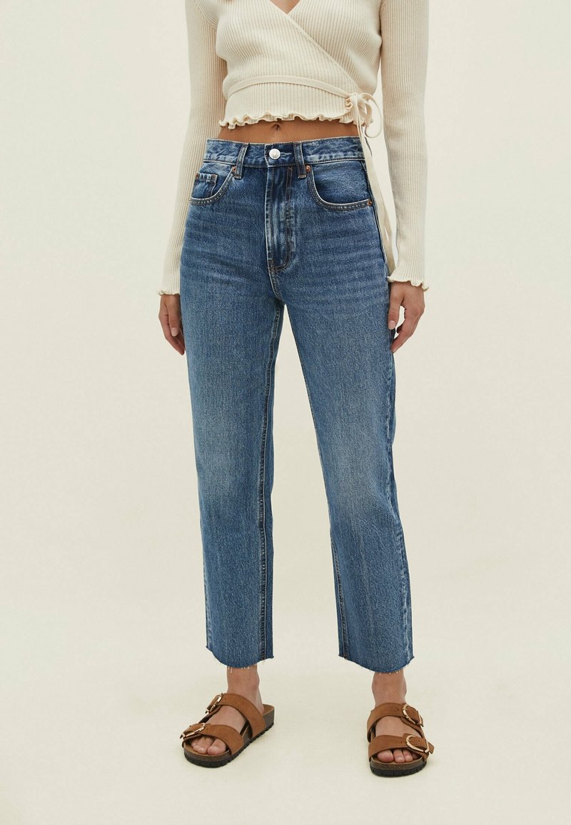 Femme portant un jean taille haute bleu, un crop top crème croisé, et des sandales marron à boucle, debout devant un fond clair uni.