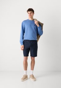 Lys blå langermet genser med en liten logo, kombinert med marineblå shorts og beige sneakers. Modellen holder en beige jakke over den ene skulderen.