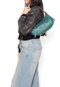 Borsa a tracolla in pelle verde acqua con forma arrotondata, tasca frontale con zip e dettagli a borchie, abbinata a una giacca nera e jeans chiari.