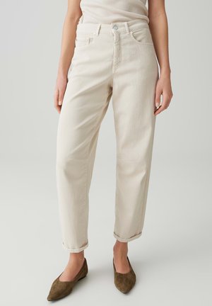 Vrouw draagt off-white rechte pijpen broek met opgerolde boorden en bruine puntige platte schoenen, staand tegen een effen lichte achtergrond.