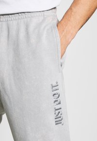 Ljusgrå träningsbyxor gjorda av mjukt tyg, med resårmidja och sidofickor. "Just Do It"-logotyp i metallisk silver på benet.