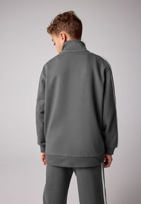 Siva jopa z visokim ovratnikom, rebraste teksture in belimi stranskimi črtami. Nošena z usklajenimi hlačami, prikazana od zadaj.