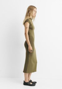 Olívovo zelené priliehavé šaty s krátkymi rukávmi, až po kolená, hladká textúra. Nosené s čiernymi topánkami s detailmi na remienku.