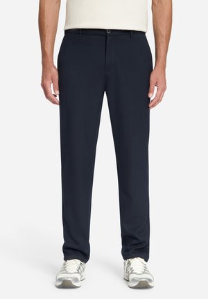 Man draagt donkerblauwe, rechte broek met riemlussen, knoopsluiting en witte sneakers, staande tegen een effen achtergrond.