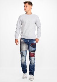 Grijze sweatshirt met lange mouwen en een ronde hals, gecombineerd met blauwe patchwork jeans met rode en zwarte accenten en contrasterende stiksels.