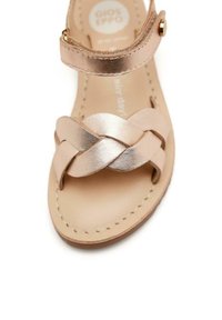 Sandalia de cuero en color oro rosa con diseño de tiras trenzadas, hebilla en el tobillo y plantilla lisa en beige. Suela plana con mínimos detalles de costura.