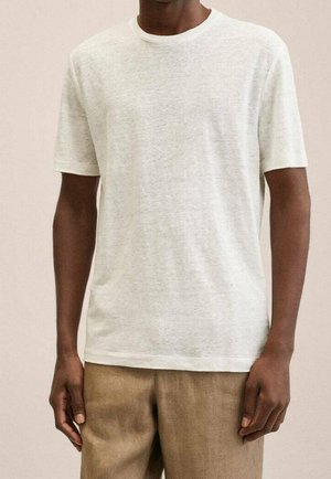 Mand iført en ensfarvet hvid kortærmet T-shirt og lysebrune bukser, stående foran en neutral beige baggrund.