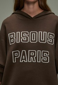 Felpa marrone con una vestibilità rilassata, caratterizzata da testo ricamato bianco "BISOUS PARIS". Il tessuto appare morbido con una consistenza moderata e un cappuccio.