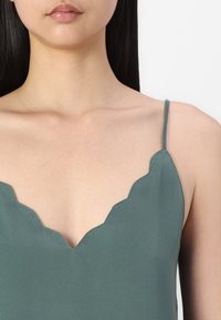 Camisole en satin vert doux avec un col festonné et des bretelles fines. Présente une texture lisse et un design simple et élégant.