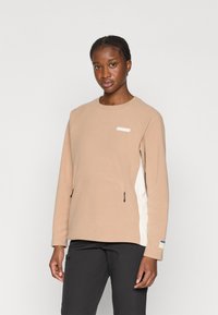 Vaaleanruskea fleece-huppari, jossa on teksturoitu pinta, valkoiset sivupaneelit ja vetoketjutaskut. Edessä pieni logo rintapielellä.