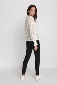 White lace top with floral pattern and ruffled cuffs, paired with black skinny jeans and white sneakers. Model shown from the back.