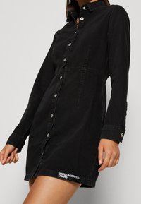 Černé džínsov�é šaty s dlouhými rukávy, límcem, předními knoflíky a bočními kapsami. Obsahují logo na lemu. Přiléhavý střih a hladká textura.