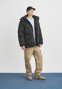 Fekete puffdzseki kapucnival, gomb- és cipzárzárral, világoskék kötött pulóver fölött viselve. Bézs cargo nadrággal és szürke csizmákkal párosítva.