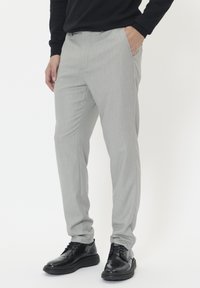 Man wearing light gray slim-fit pants with hand in pocket and black dress shoes, standing against a white background.