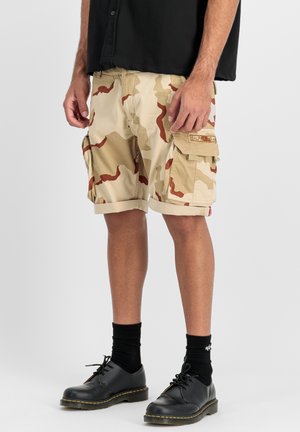 Camouflage cargo shorts in beige with red accents, featuring large side pockets and a rolled hem. Worn with black shoes and socks.