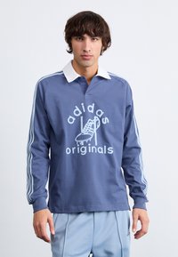 Polo majica dugih rukava plave boje s bijelim ovratnikom i tri bijele pruge na rukavima. Sadrži grafički otisak tenisice i natpis "adidas originals."