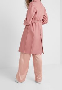 Růžový kabát z vlněné směsi s páskem v pase, dlouhými rukávy a bočními rozparky, doplněný lesklými světle růžovými kalhotami s širokými nohavicemi a bílými botami.