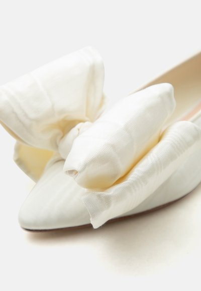 Chaussure plate couleur crème à bout pointu avec un grand nœud en tissu doux sur le devant, présentée sur fond blanc.