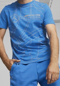 Blå kortärmad t-shirt med ett geometriskt vitt mönster och logotyper för AMG och Petronas, tillverkad av mjukt bomullstyg.