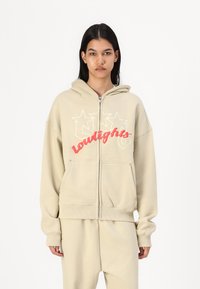 Beige zip-up hoodie with a large red and white "lowlights" graphic, featuring a front pocket and soft, textured fabric.