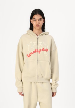 Šviesiai rudos zip-up gobtuvė su dideliu raudonu ir baltu "lowlights" grafikos atvaizdu, turinti priekini pocket'ą ir minkštą, tekstūruotą audinį.
