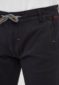 Dark gray pants made of textured fabric, featuring a woven black and white drawstring, yellow stitching accents, and a small brand tag.